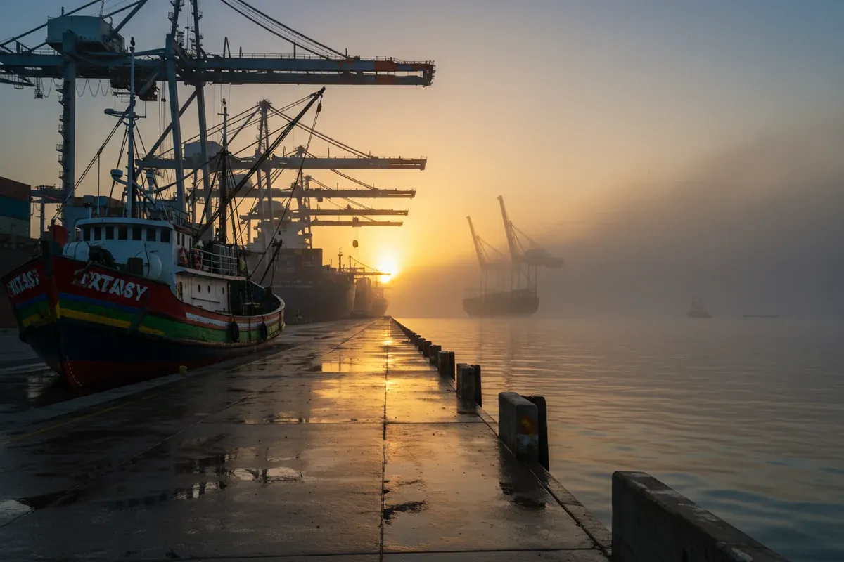 A weathered concrete pier stretches into a hazy distance showcasing large container ships unloading cargo at Americas