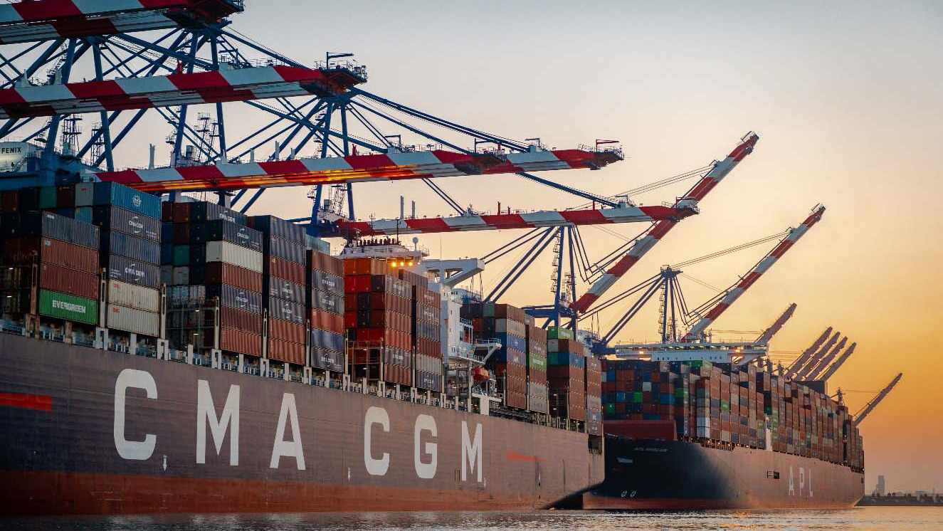 A large container ship CMA CGM floats in the ocean with towering cranes in the background