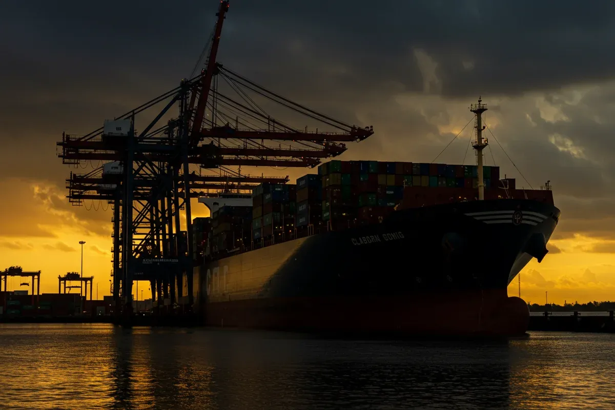 A dark ship silhouette sits in a port with towering cranes against a dusky sky emphasizing the industrial scene