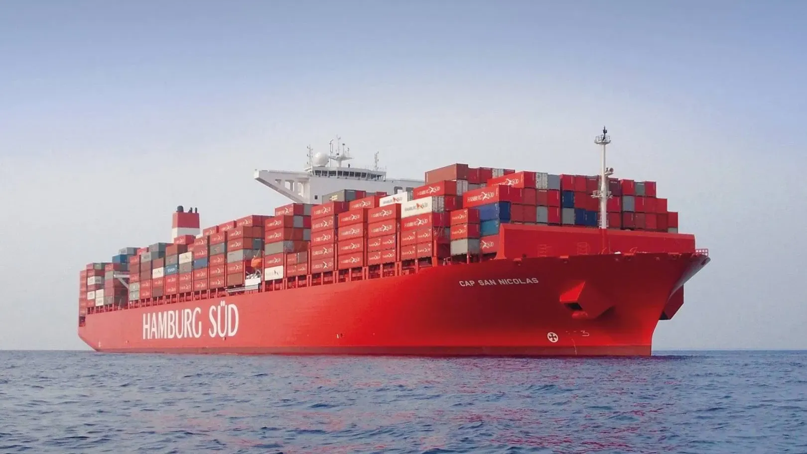 A large red container ship named Hamburg Süd sails on a calm ocean with a prominent ships mast and visible cargo containers