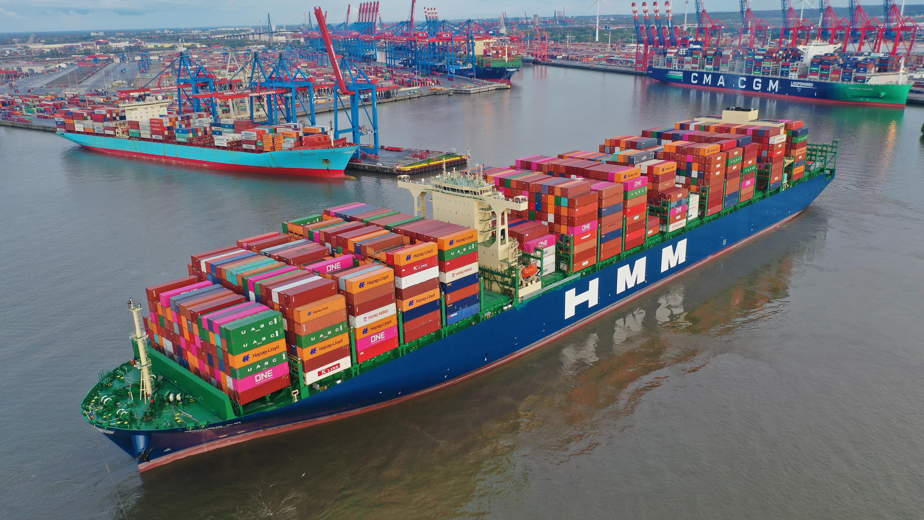 A large container ship dominates the image with a prominent CMA CGM name and numerous shipping containers visible onboard