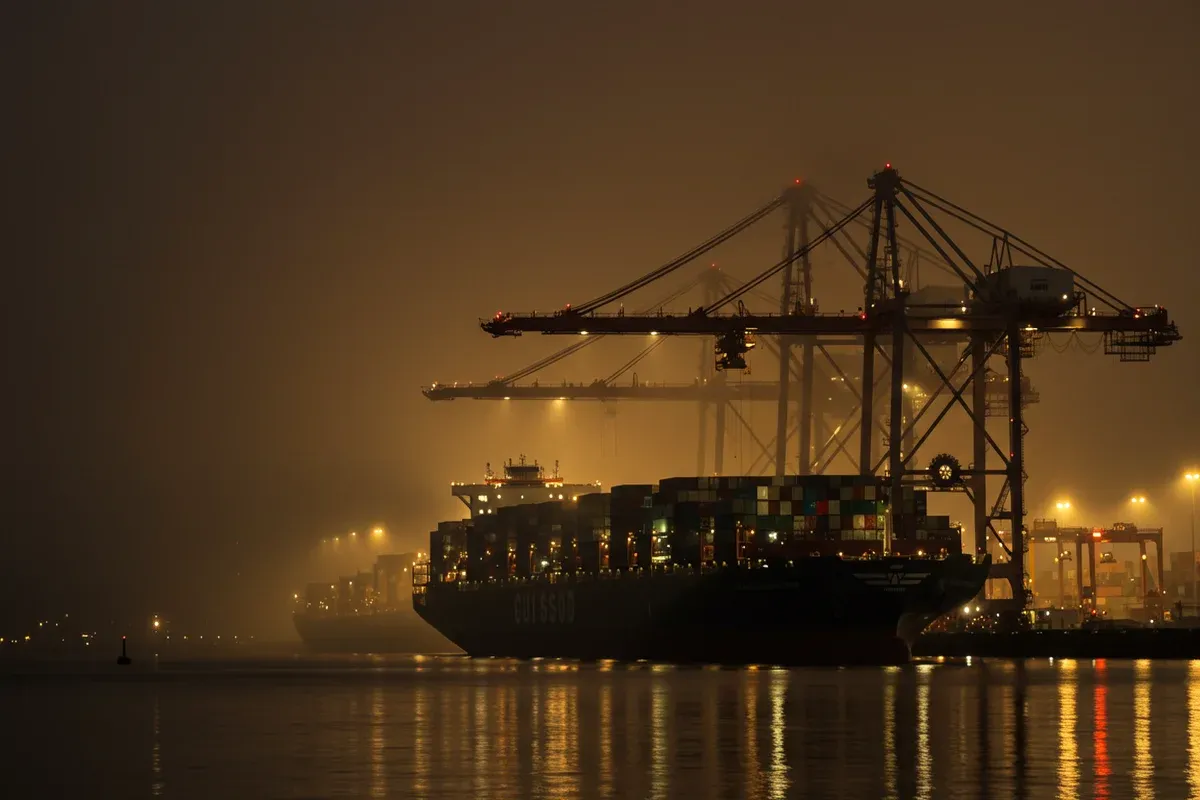 A large container ship navigates a waterway under towering cranes reflecting harbor lights in the murky water