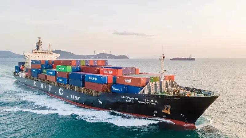 A large container ship navigates the ocean under a cloudy sky with visible cargo containers onboard