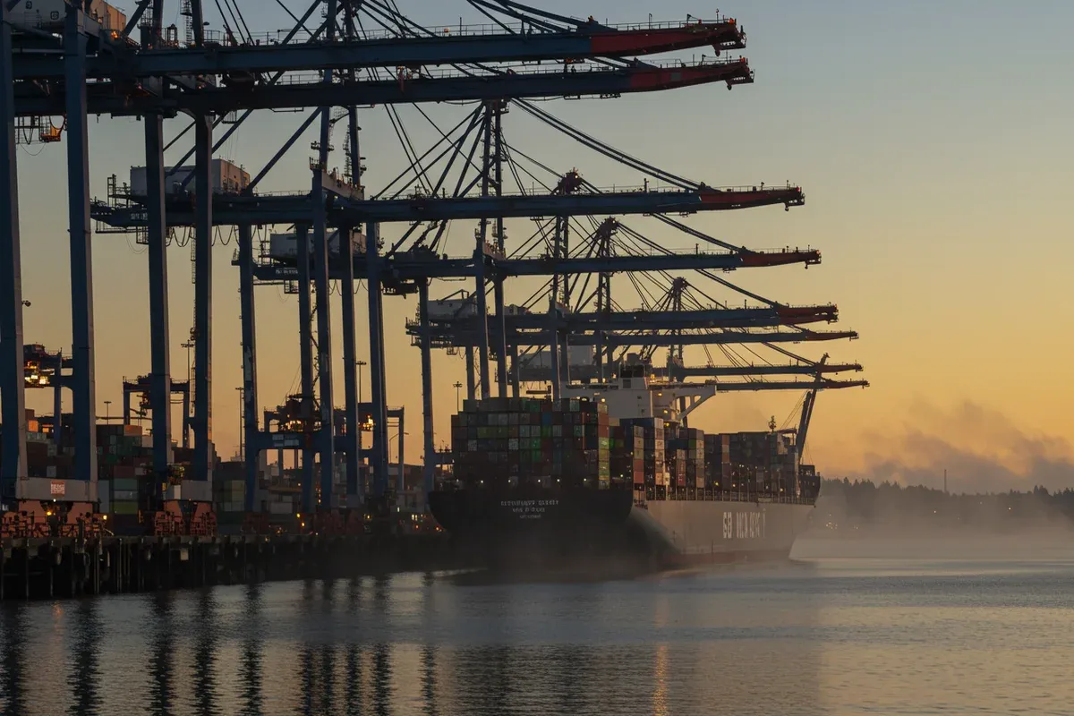 Vessels navigate the Puget Sound under towering cranes at the Northwest Seaport Alliance docks reflecting in the calm water