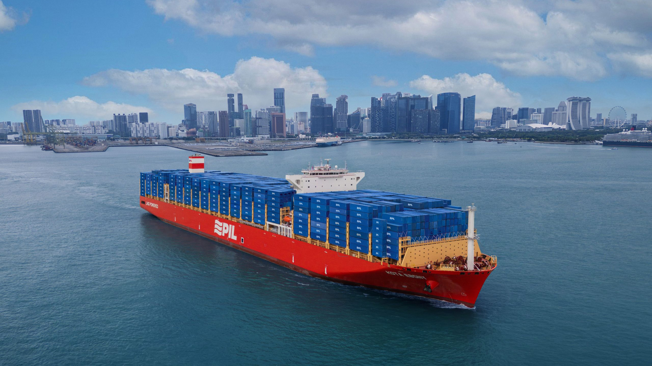 A large red container ship the Pacific International Lines vessel navigates the harbor against a dense cityscape