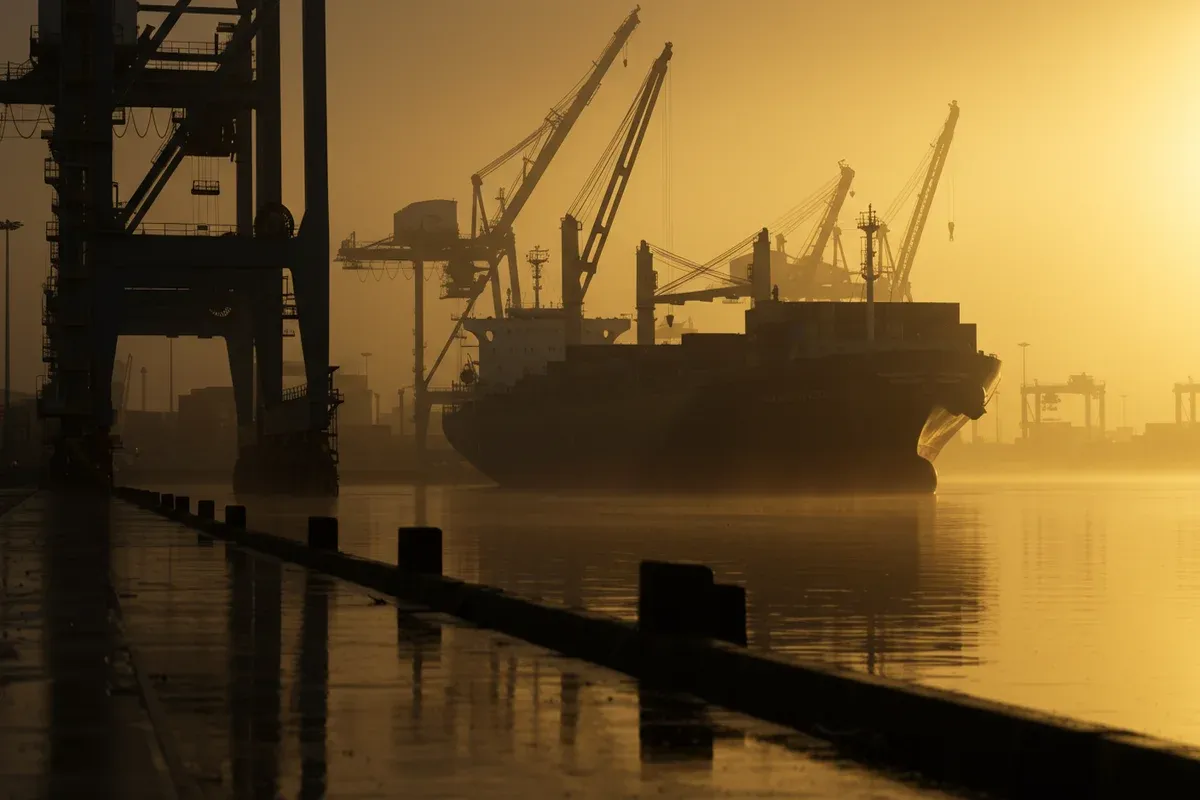 A large cargo ship is silhouetted against a misty harbor background with prominent cranes and a waterfront rail