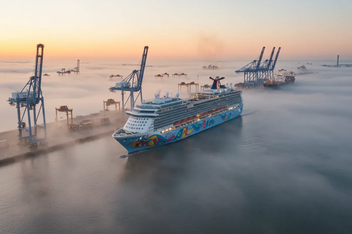 A brightly colored cruise ship approaches a busy port filled with towering cranes and industrial structures