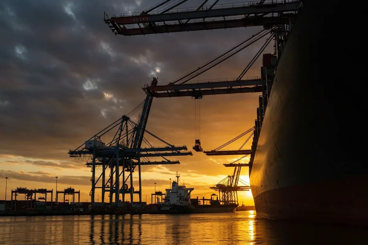 A large ship sits in the harbor reflecting the warm golden light of sunset illuminating cranes and docks along the water