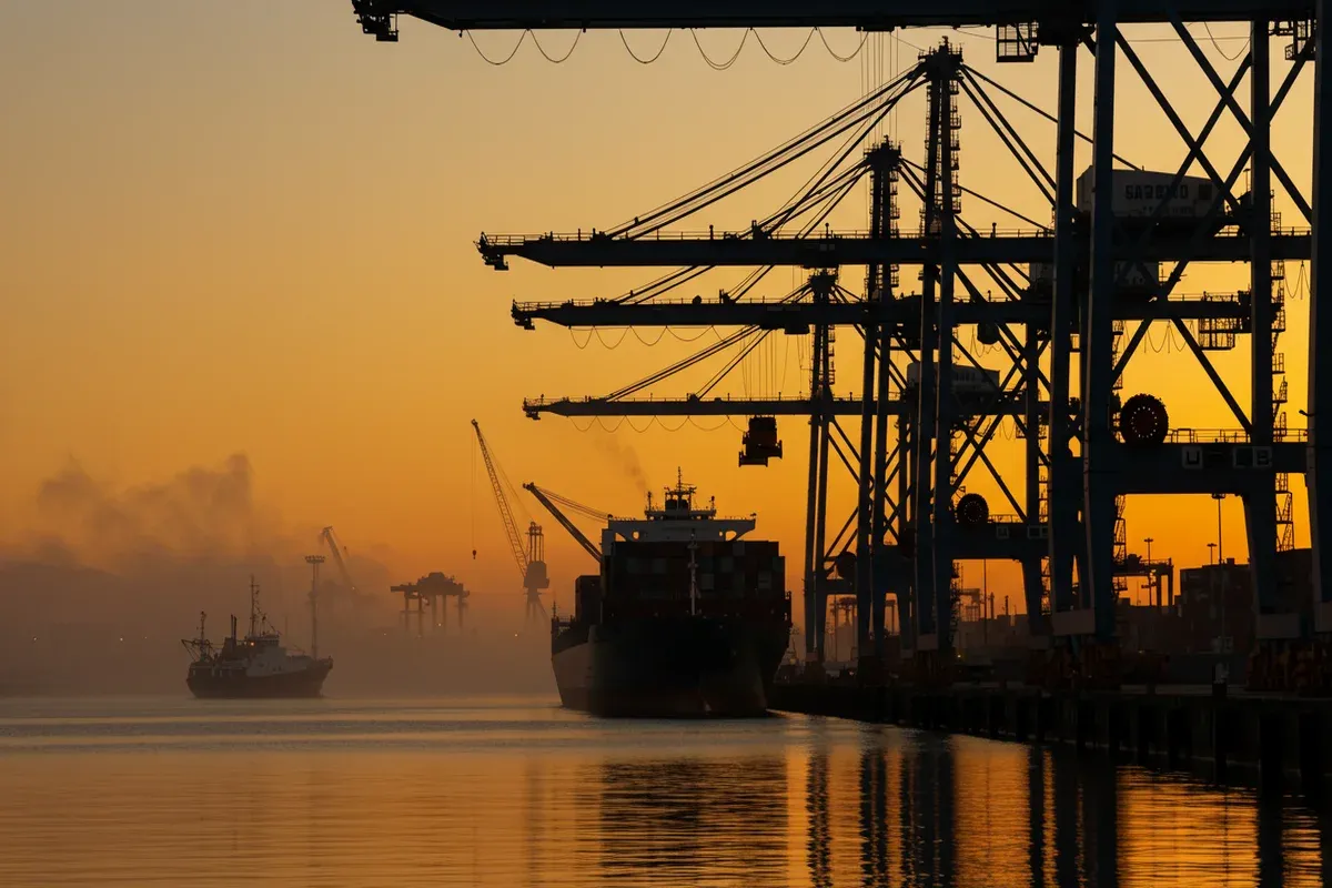 A container ship navigates a misty harbor filled with towering cranes and ships reflecting in the calm water