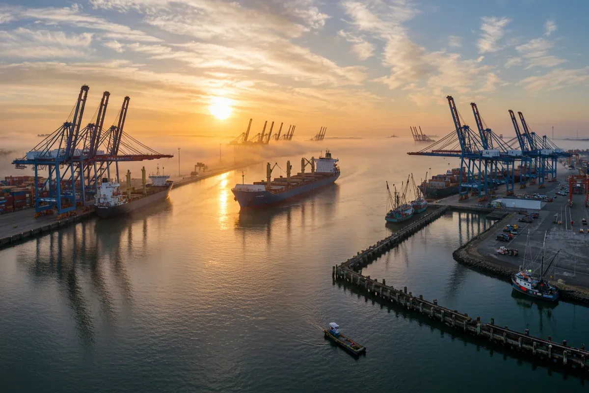 A large cargo ship navigates a busy port channel with docks and a small boat alongside a concrete seawall during sunrise