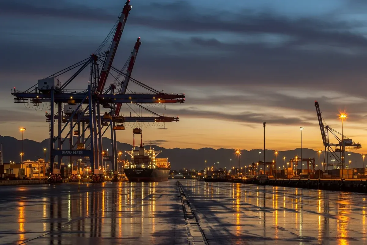A long pier extends into a harbor filled with towering cranes reflecting city lights creating a dynamic scene of maritime