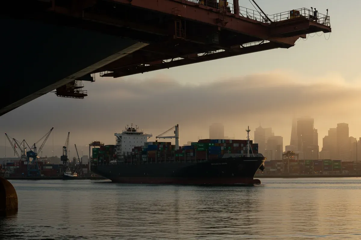 A large container ship navigates the San Francisco Bay waters under a golden sunset light reflecting on the waters surface