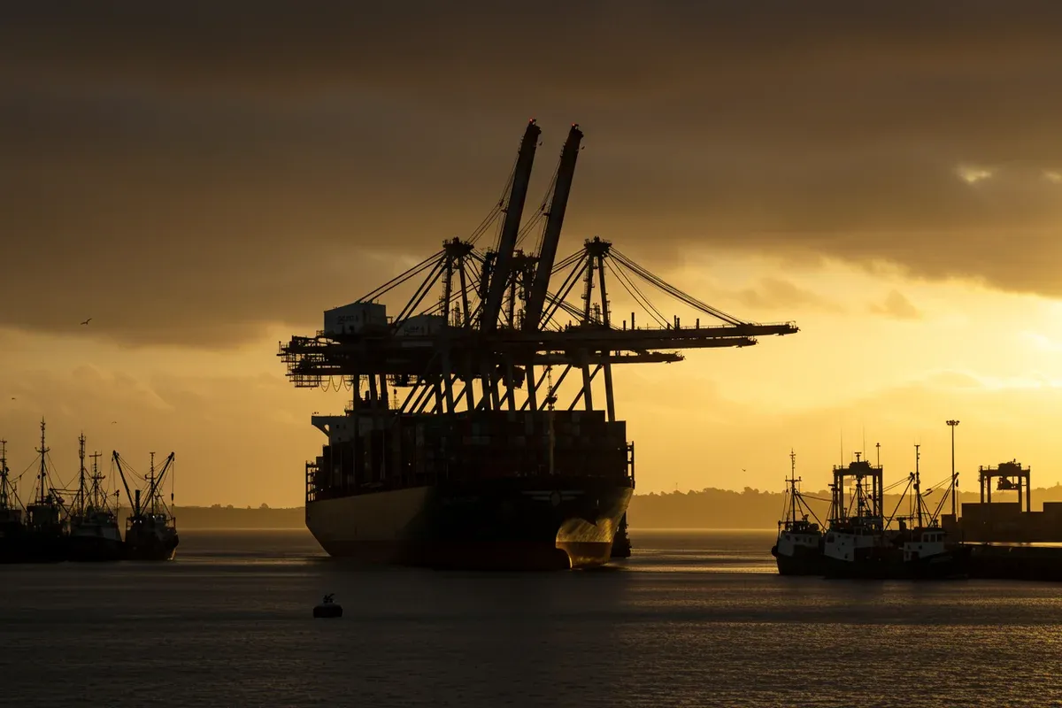 A large container ship navigates the Port of St Helens with numerous fishing boats dotting the calm waters