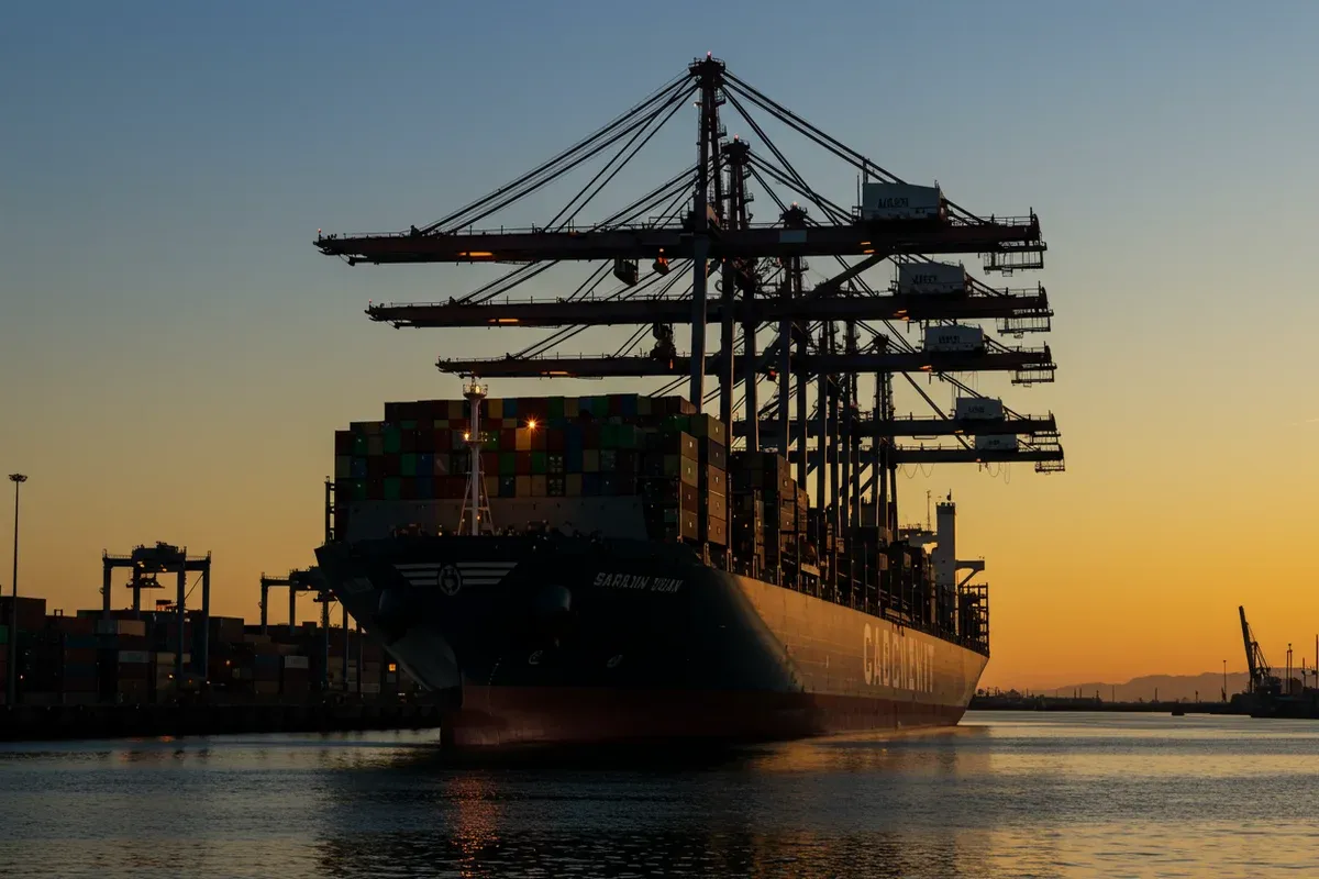 A large cargo ship navigates the Sacramento River amidst towering cranes and maritime port infrastructure