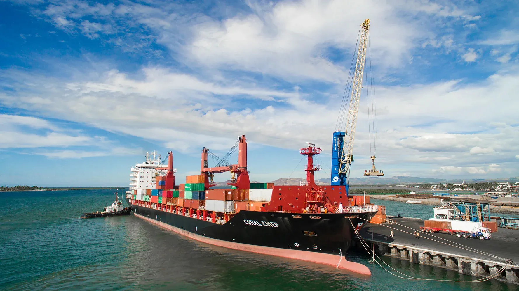 A large container ship the Swire Shipping vessel navigates a harbor channel alongside a pier with cranes in the background