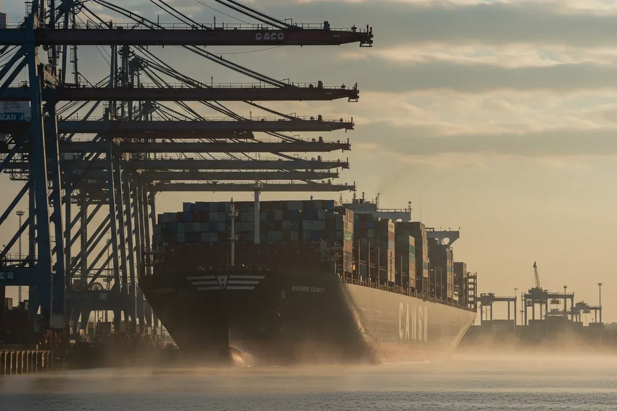 A large cargo ship dominates the scene at Vicksburg Port with towering cranes extending into the hazy distance
