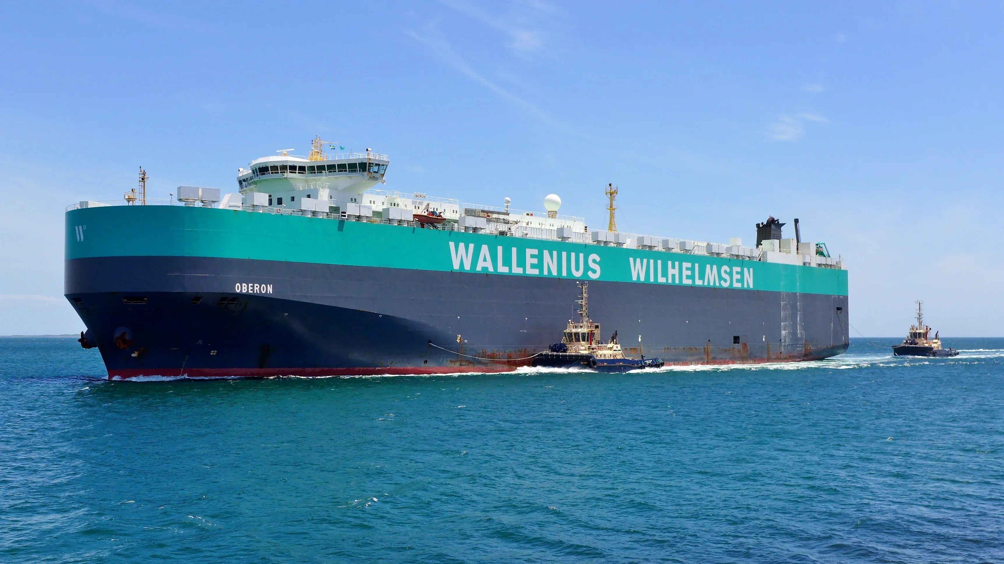 A large container ship the Wallenius Wilhelmsen navigates the ocean with two tugboats assisting its movement