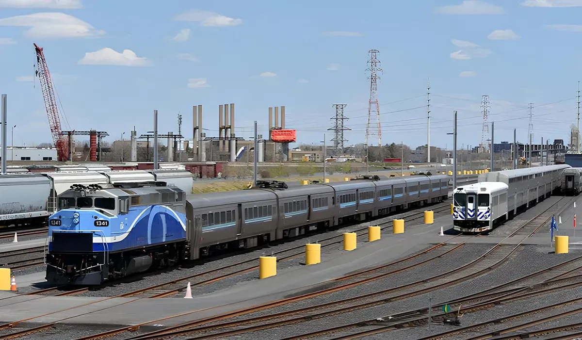 A blue Amtrak train sits on a railway track with yellow safety barriers visible in the background near a large industrial