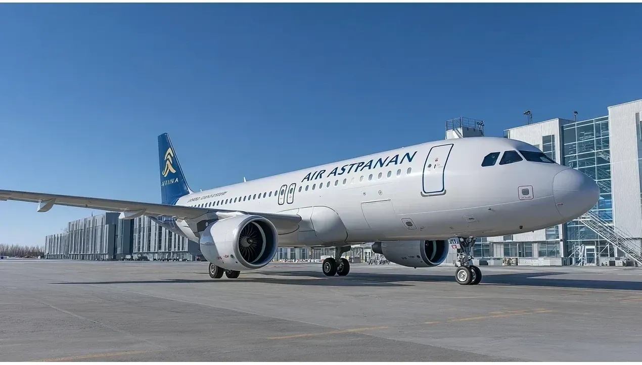 A white airplane of Air Astana is prominently displayed on a light gray tarmac with modern buildings in the background