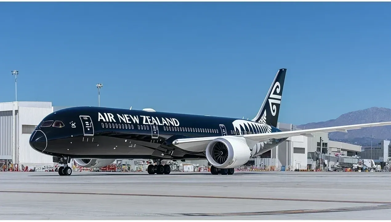 A large Air New Zealand aircraft sits on a tarmac near airport buildings reflecting sunlight revealing its distinctive