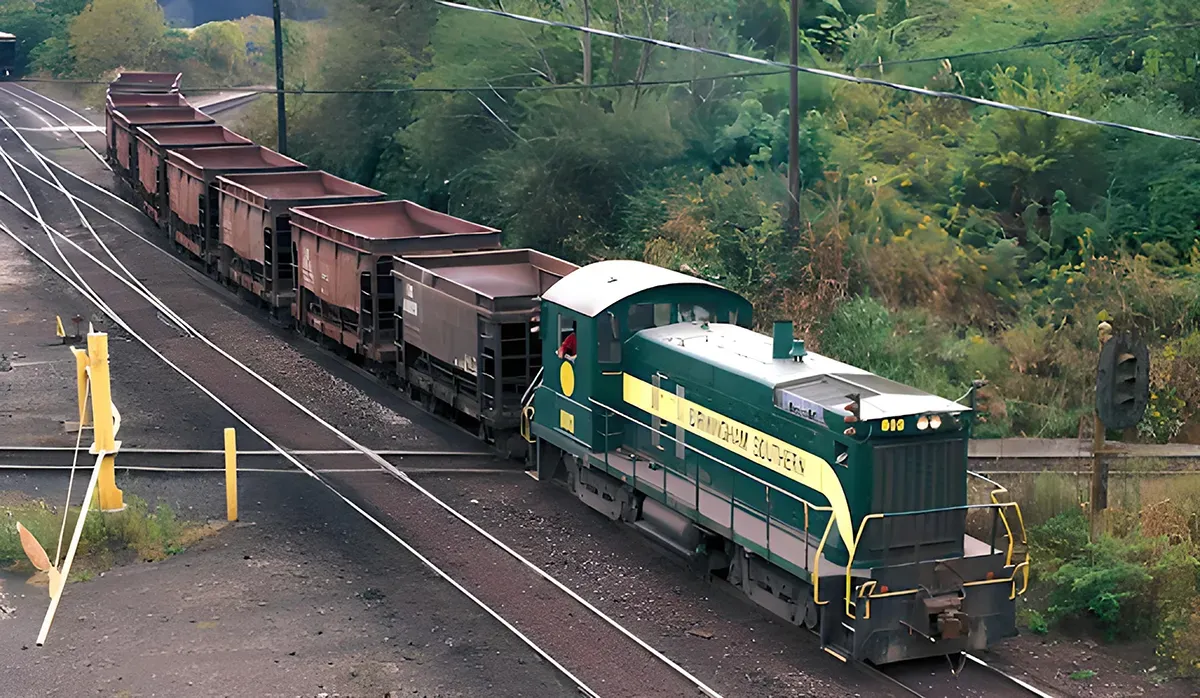 A green Birmingham Southern Railroad efficient supply chain locomotive sits on a rail line with yellow warning signs in the