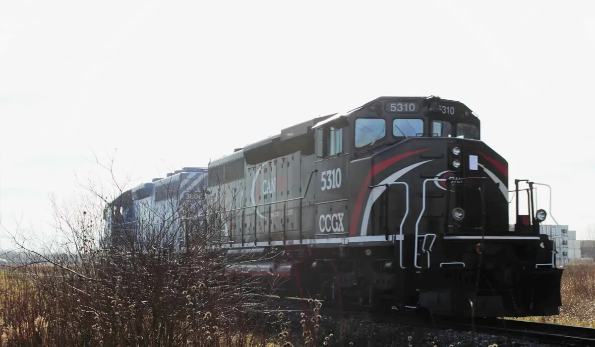 A freight train moves steadily along a track through a barren landscape with dried grasses and a distant skyline suggesting