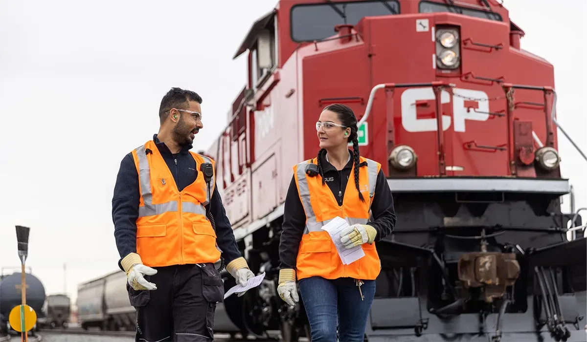 Two workers in orange vests walk along a railway track inspecting a long freight train