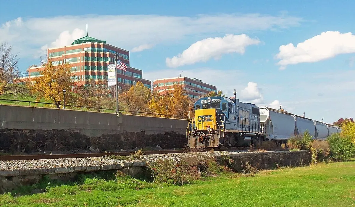 A CSX Transportation freight train travels along a trackside embankment with a concrete retaining wall and buildings in the