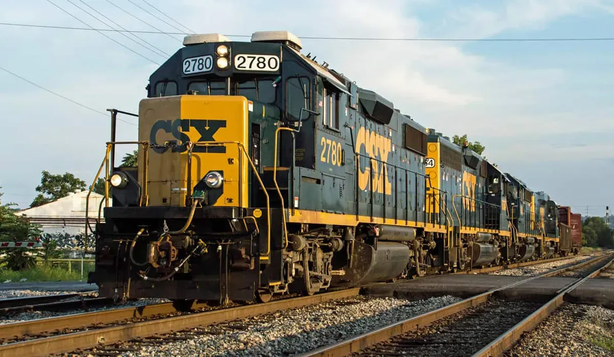 A CSX freight train travels along a gravel track with a long boxcar and engine visible in the foreground