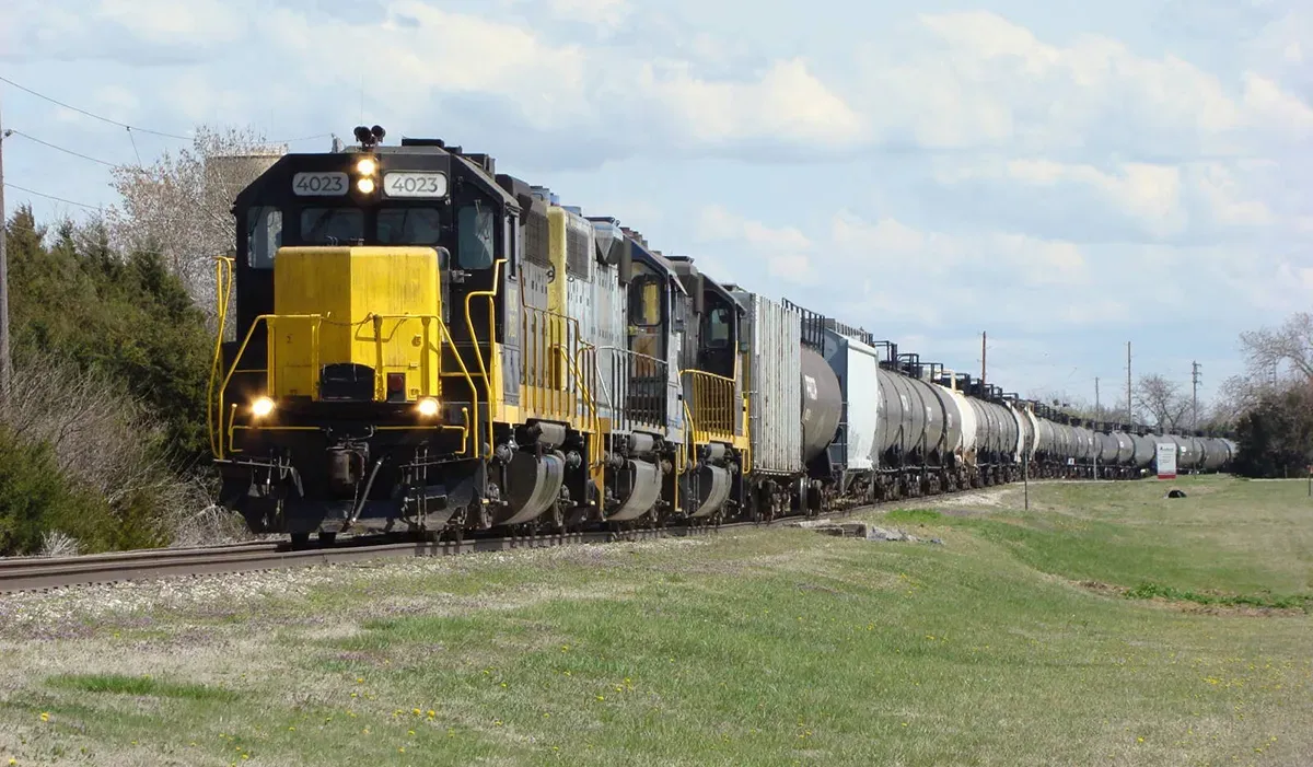 A long freight train extends across a grassy field with several boxcars in a consistent black and yellow color scheme moving