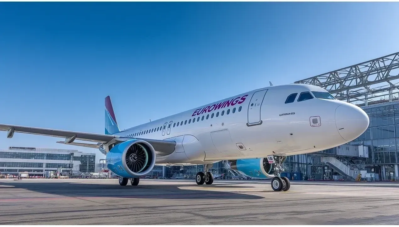 A bright Eurowings aircraft stands on the tarmac at an airport terminal with modern architecture visible in the background