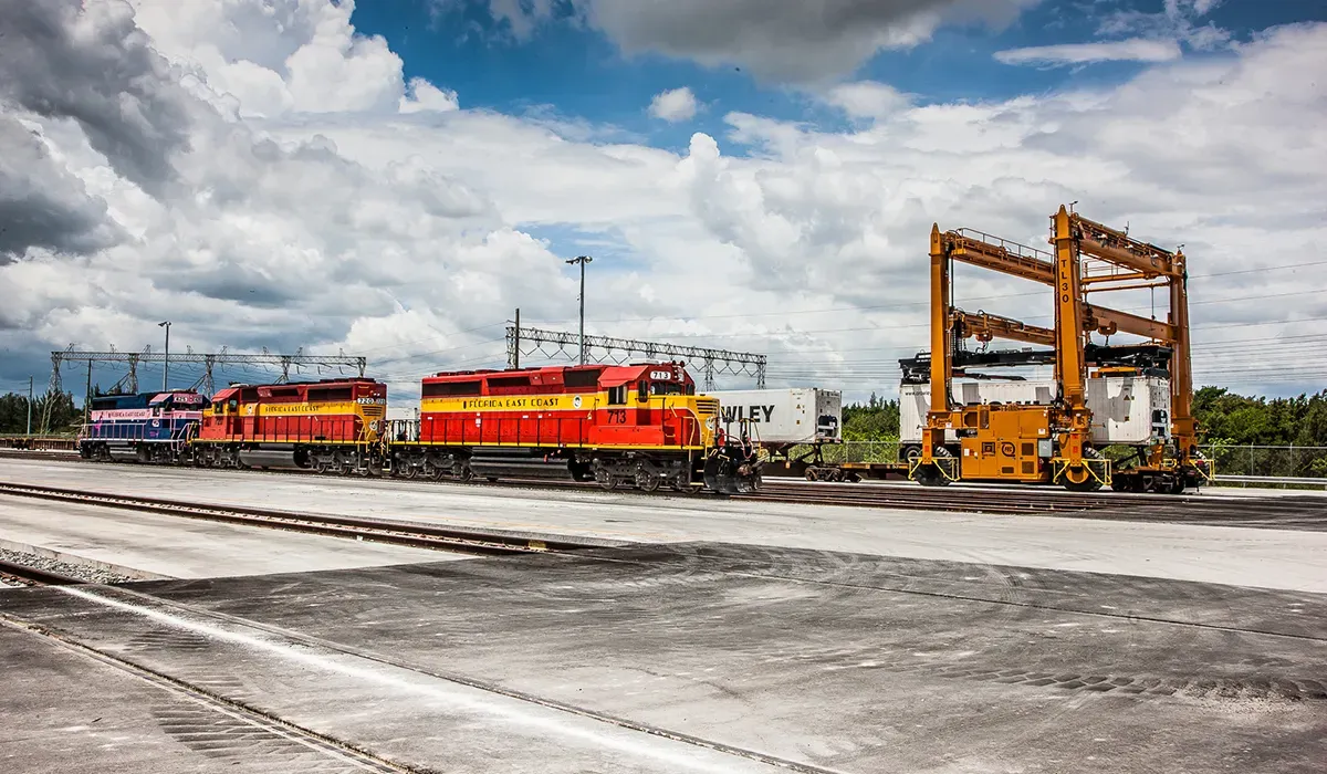 A freight train sits on a concrete track with large orange cranes in the background signifying efficient supply chain