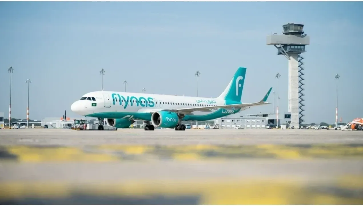 A bright white Boeing 737 aircraft of flynas stands on the tarmac with a tall control tower in the background