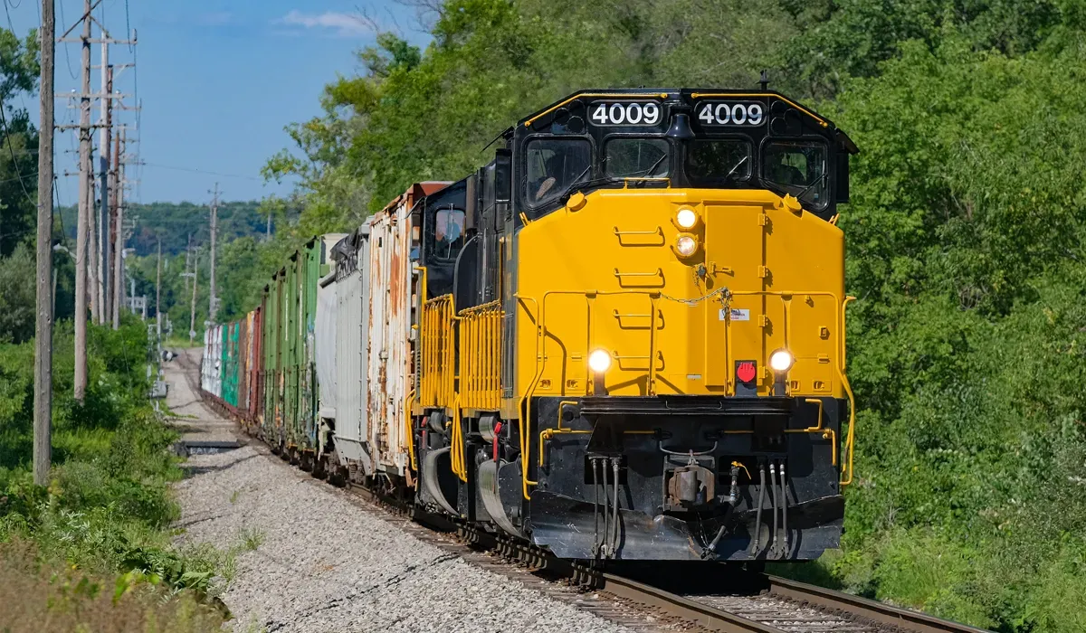 A yellow freight train travels down a rural track with multiple trees lining the background The train appears efficient and