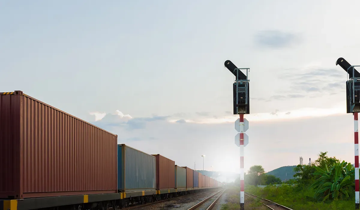 Rows of red freight cars stretch along a railroad track in an industrial area with a hazy sky Signal lights stand tall above