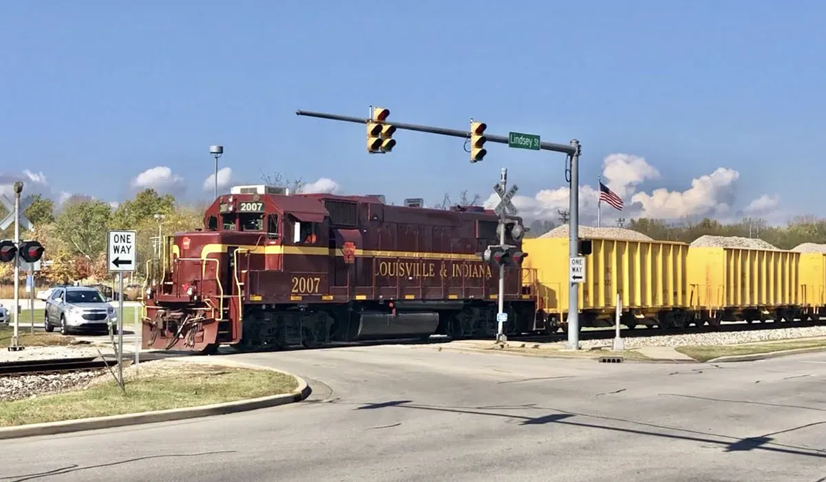 A Louisville and Indiana Railroad locomotive pulls a freight train along a roadside with a green stop sign and city street