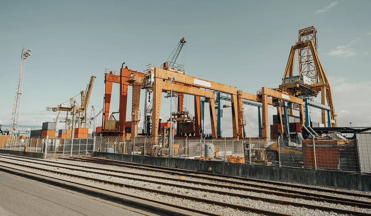 Orange cranes stand alongside a railway line and chainlink fence at a shipping terminal highlighting efficient supply chain