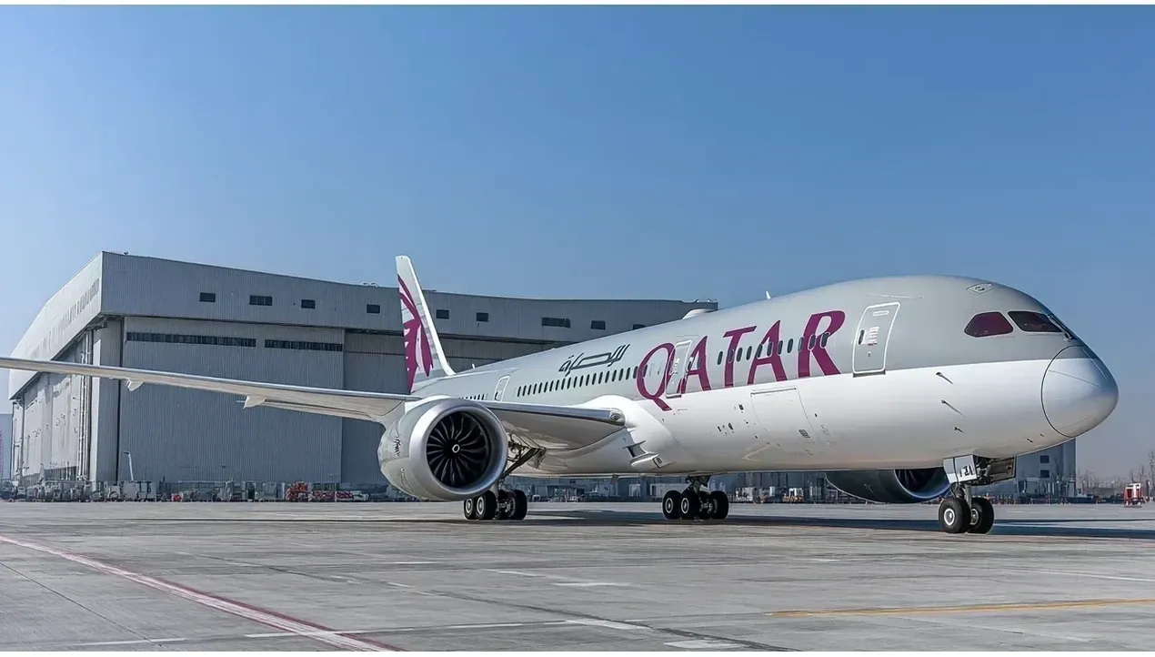 A Qatar Airways aircraft stands at an airport gate against a large industrial building highlighting its brand colors