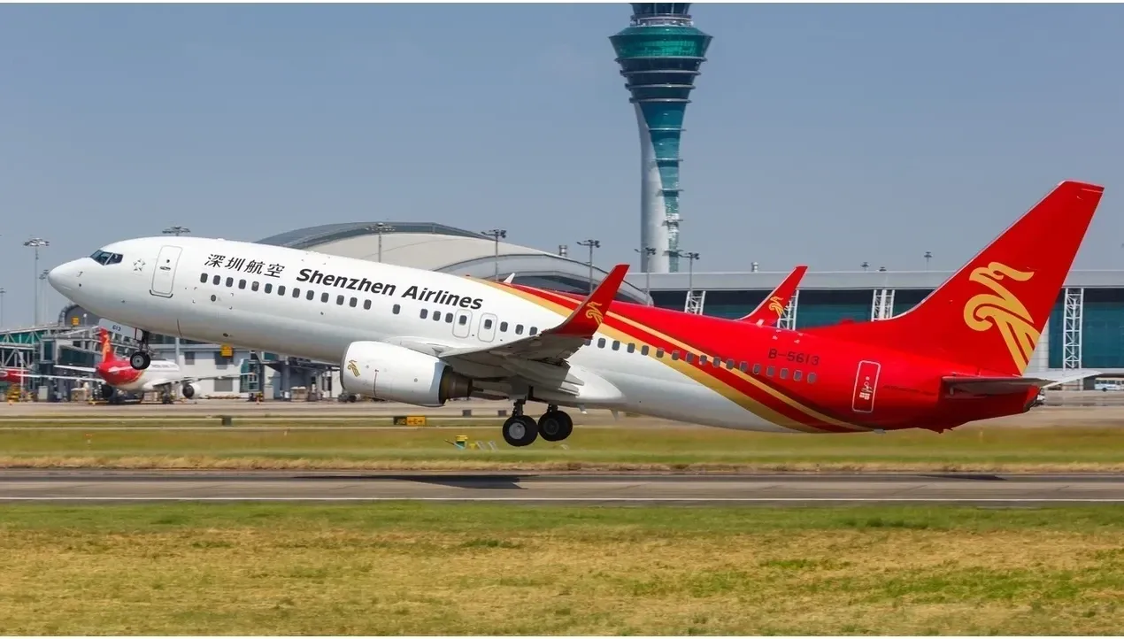 A vibrant Shenzhen Airlines Boeing 737 aircraft dominates the image soaring through the sky with its distinctive red hero