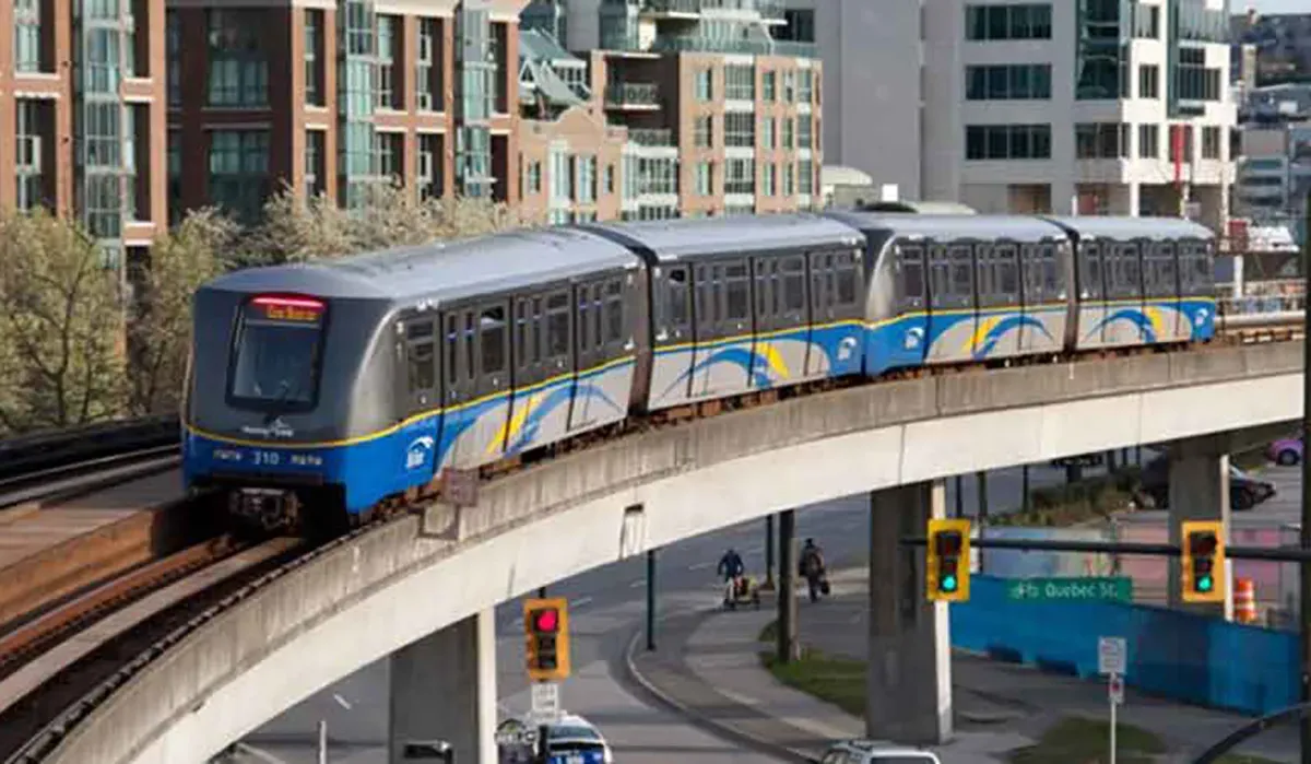 A blue SkyTrain rapid transit vehicle swiftly travels along elevated tracks beneath a modern city overpass structure