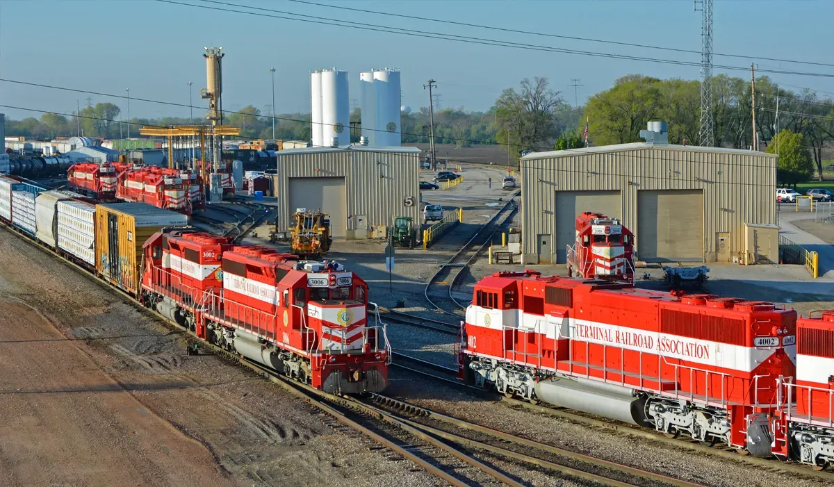 Red freight trains transport goods across a terminal yard with industrial buildings and a gravel track visible in the