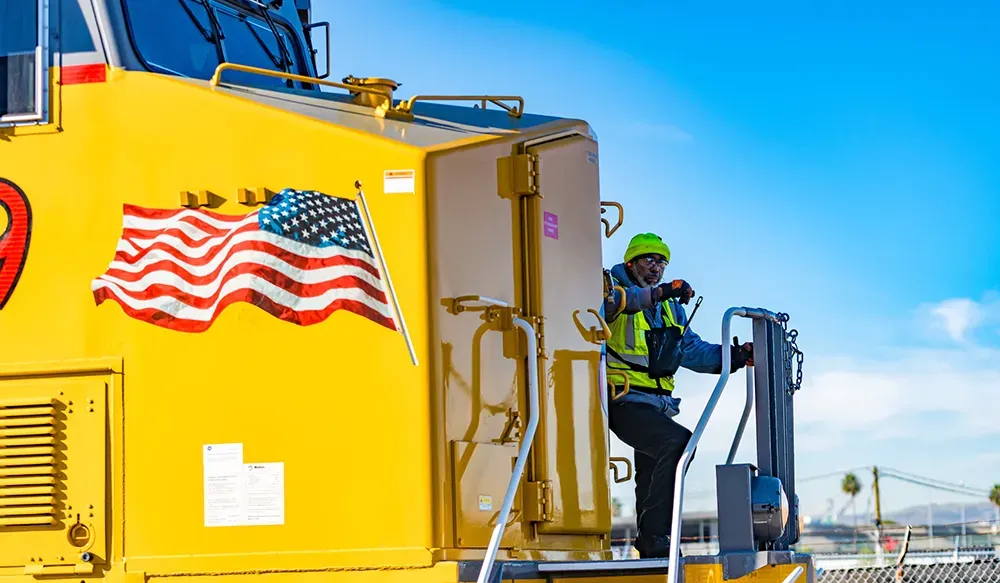 A worker in a yellow safety vest stands on a bright yellow locomotive overlooking a harbor The Union Pacific logo is