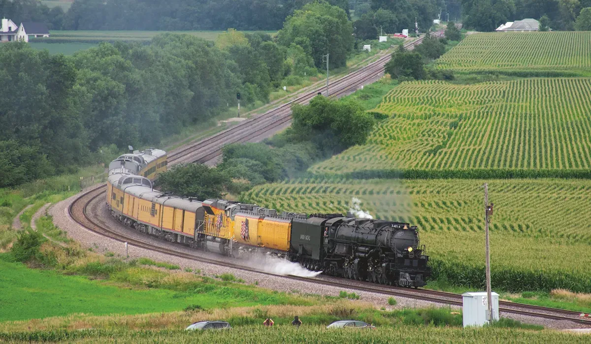 A powerful Union Pacific locomotive blasts steam as it curves around a green hillside conveying supplies