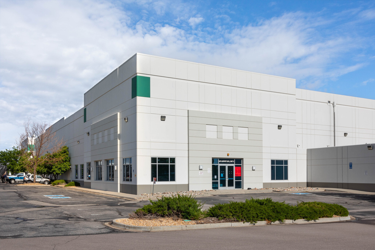 A modern warehouse building with multiple doors and large windows sits on a paved lot adjacent to a grassy area