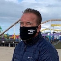 Arnold Schwarzenegger stands with a dark blue face mask against a vibrant backdrop of carnival rides and colorful lights