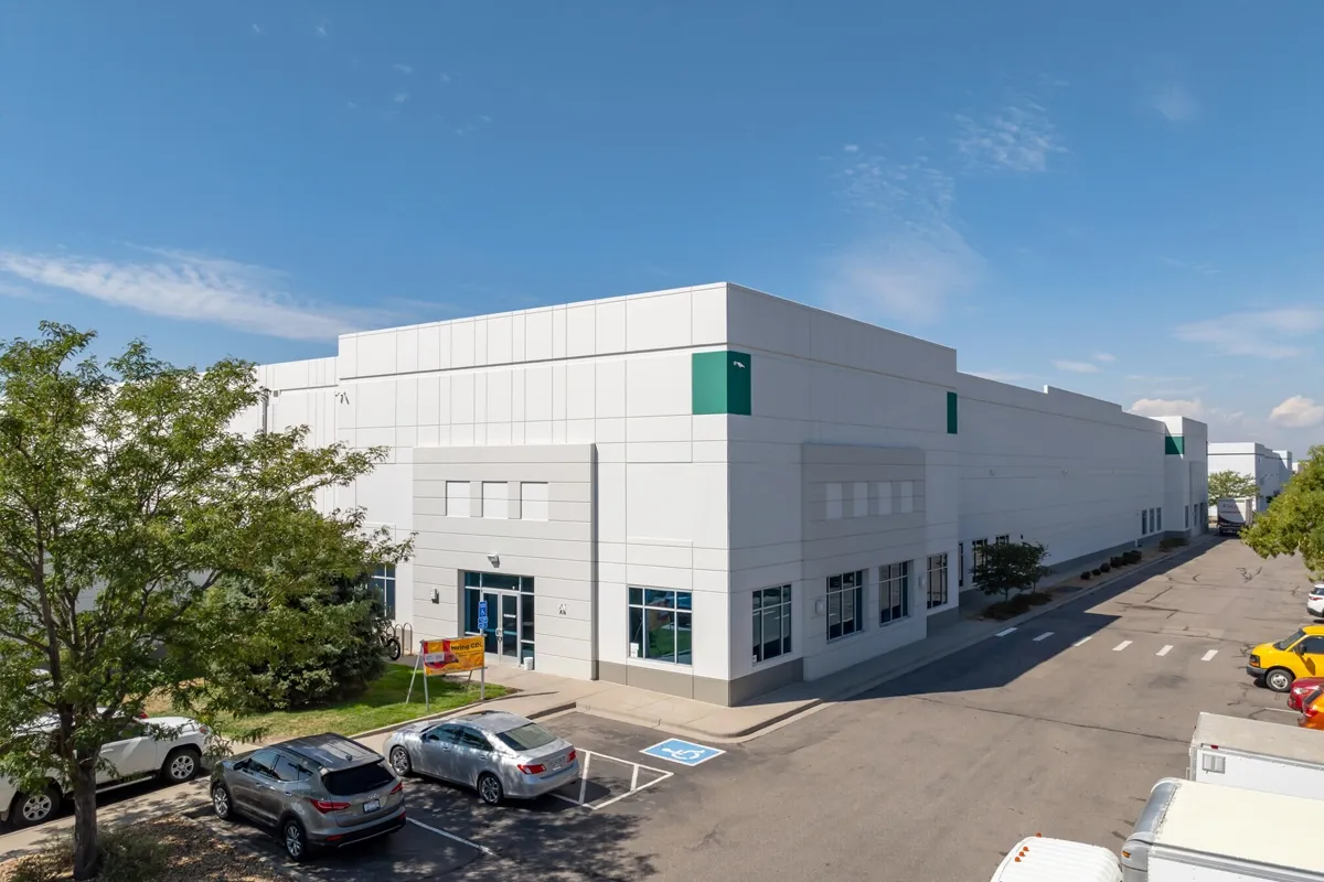 A modern industrial building complex with white walls and large windows sits on a paved road surrounded by trees and vehicles