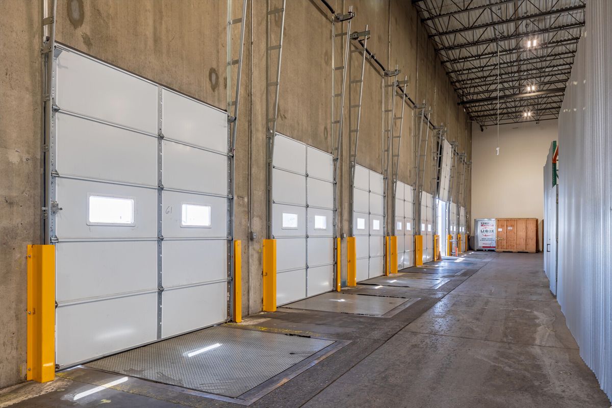 A long interior warehouse features multiple steel rollup doors and concrete floors with yellow safety barriers along the