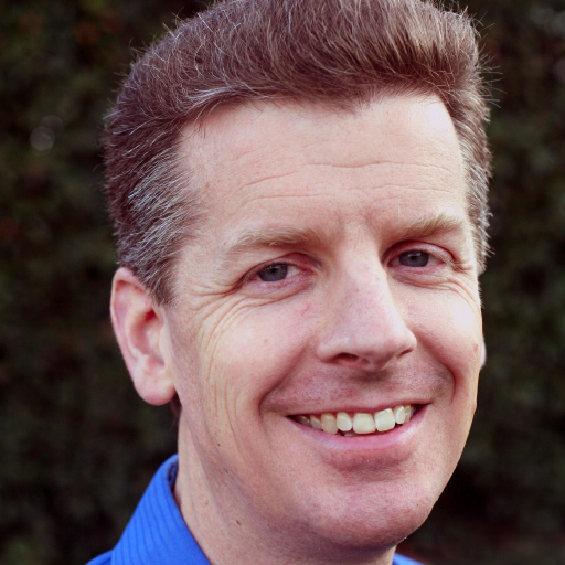 A smiling man in a blue shirt stands against a green leafy background presenting a friendly professional demeanor
