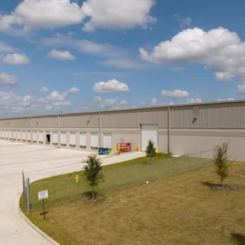 Large beige warehouse with white loading docks, landscaped lawn with evergreen trees, under partly cloudy sky