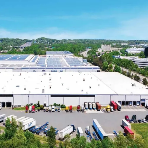 A large industrial warehouse dominates the scene with a sprawling white roof and multiple delivery trucks parked in the lot